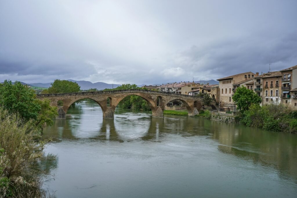 Cómo funciona Historic Puente La Reina bridge spans calm river under cloudy sky.